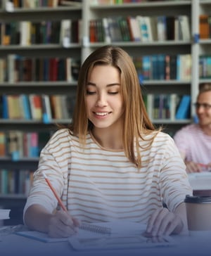 A smiling female student sitting at a library desk, writing in a spiral notebook with a pencil in front of filled bookshelves.