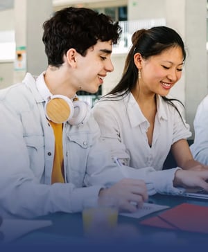 A young man with headphones around his neck taking notes in a notebook next to a smiling Asian woman during a study session.