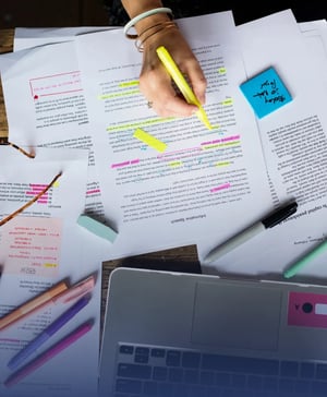 Overhead view of a student editing printed essay pages with a yellow highlighter at a desk with a laptop, sticky notes, and colored pens.